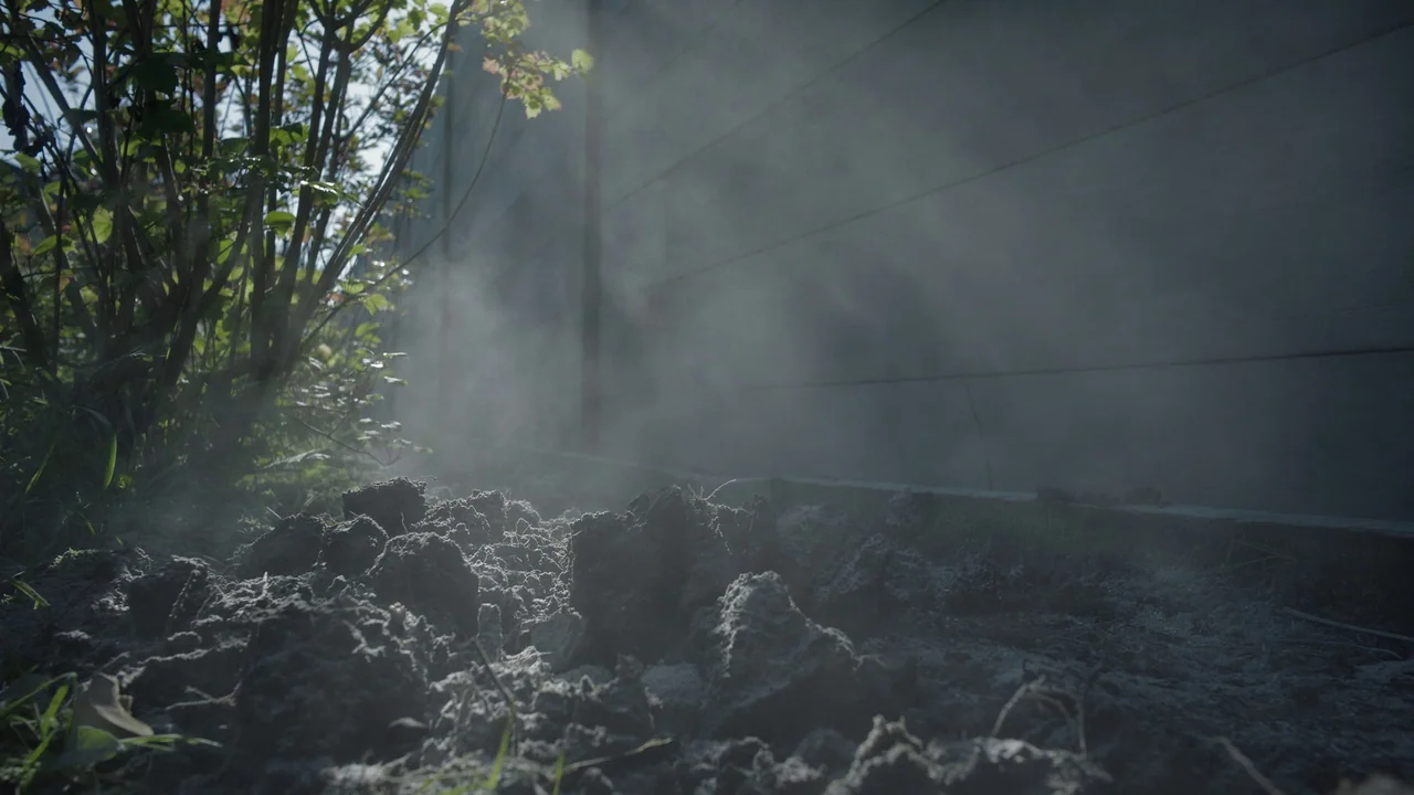 The Zone Of Interest (2023) movie still: establishing — A pile of dark soil sits next to a grey wood-plank fence. Mist rises off the damp earth…