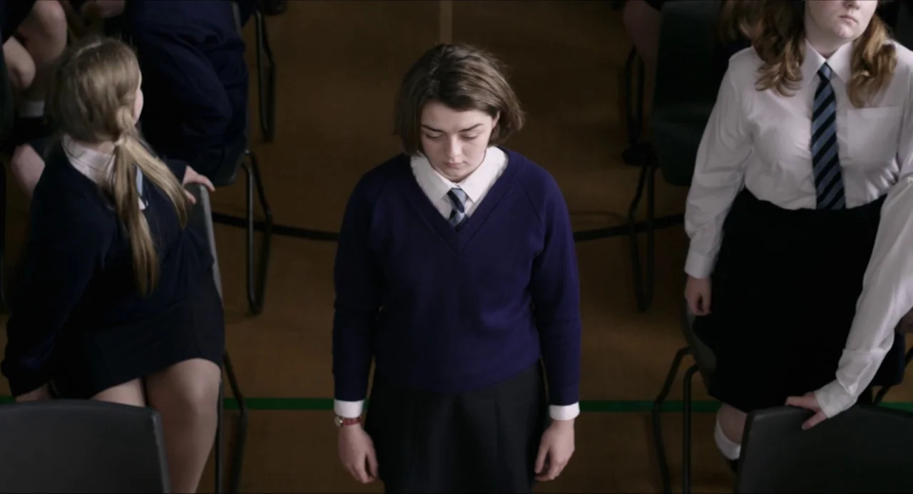 The Falling movie still: medium — A school assembly hall features rows of students sitting on black plastic chairs. A girl in a blue sweater stands…
