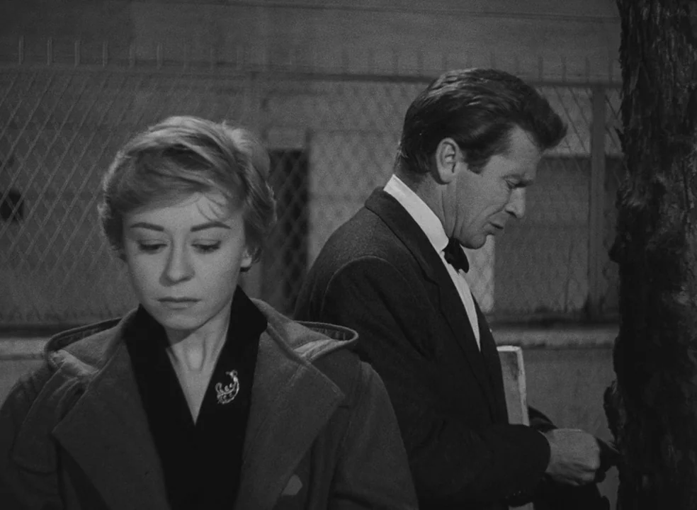 Il Bidone (1955) movie still: medium — An outdoor sidewalk at night with a chain-link fence behind the two subjects. A woman and a man stand…