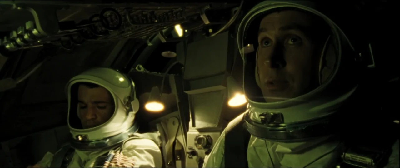 First Man (2018) movie still: medium — Two astronauts in white pressure suits are positioned inside a confined spacecraft cockpit. The men wear…