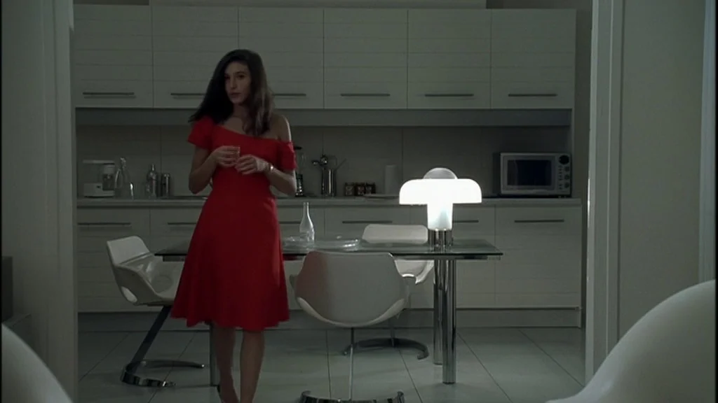 Romance (1999) movie still: fullbody — A minimalist white room features modern furniture including white chairs and a glass table. A woman in a red…