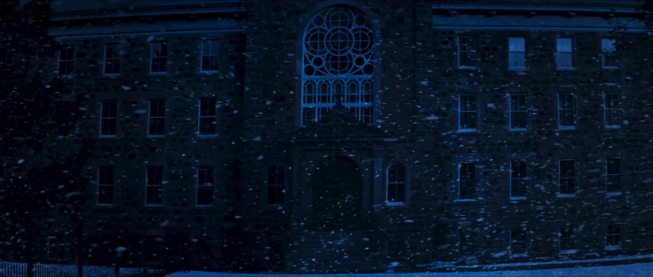 The Sacrifice Game (2023) movie still: establishing — A dark stone university building with a large arched window stands under night sky. Heavy snow…