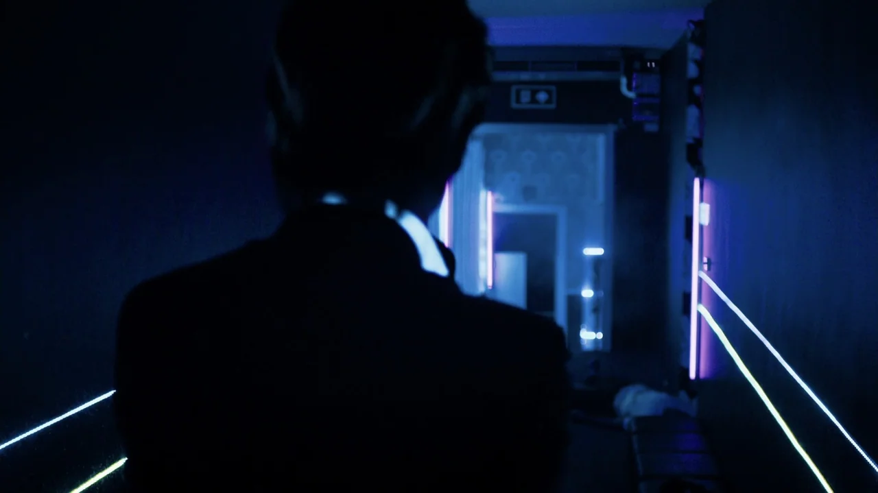 Zero Tolerance (2015) movie still: medium — A dark hallway is illuminated by thin blue neon light strips on the walls. A person in a dark suit…