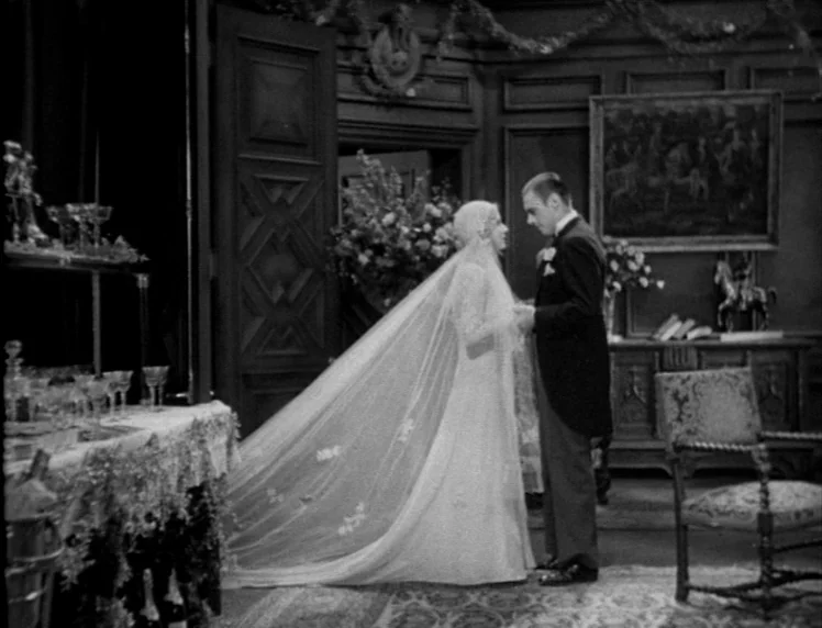 Frankenstein movie still: fullbody — A wood-paneled room decorated for a wedding with floral arrangements and champagne glasses. A bride in a long…