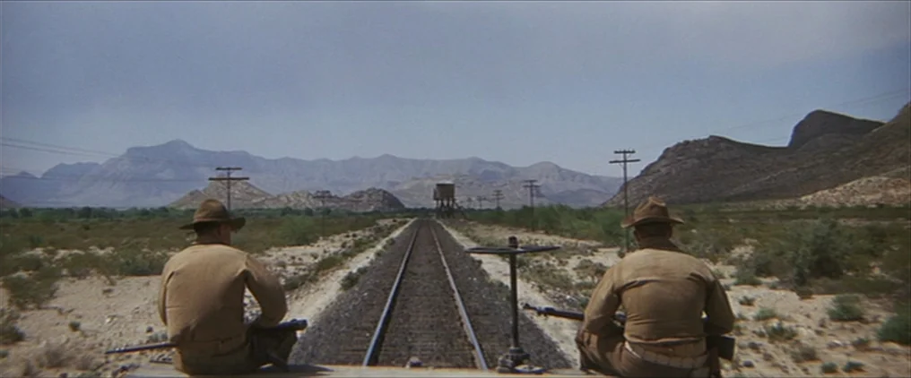 The Wild Bunch (1969) movie still: wide — An arid desert landscape features a single set of train tracks extending toward mountains. Two soldiers in…