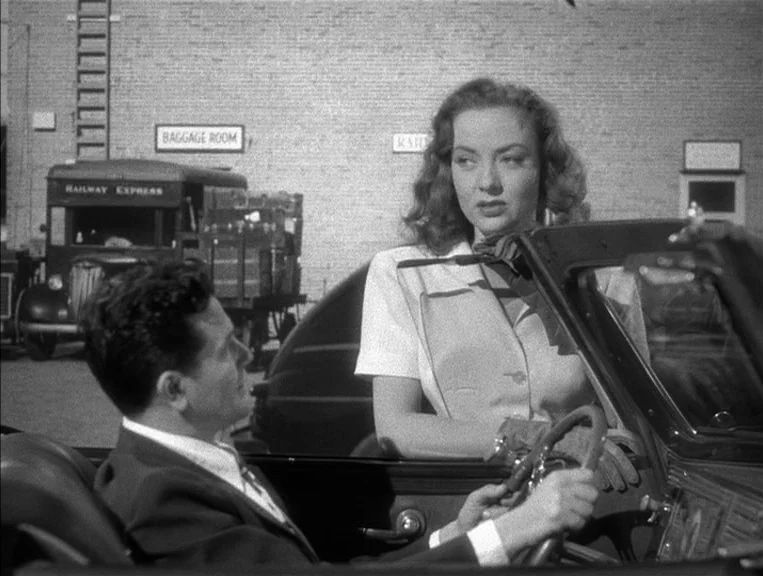 The Postman Always Rings Twice (1946) movie still: medium — A woman leans toward a man seated in a convertible parked at a station. A brick building…