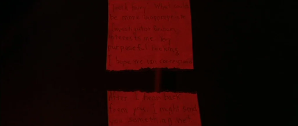 Manhunter (1986) movie still: closeup — Two scraps of red paper with dark handwritten text float against a black background. The message on the…