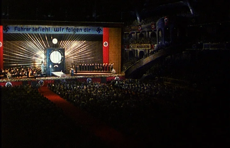 Lili Marleen (1981) movie still: wide — A dark indoor assembly hall features a grand stage with Nazi propaganda banners visible. A large crowd of…