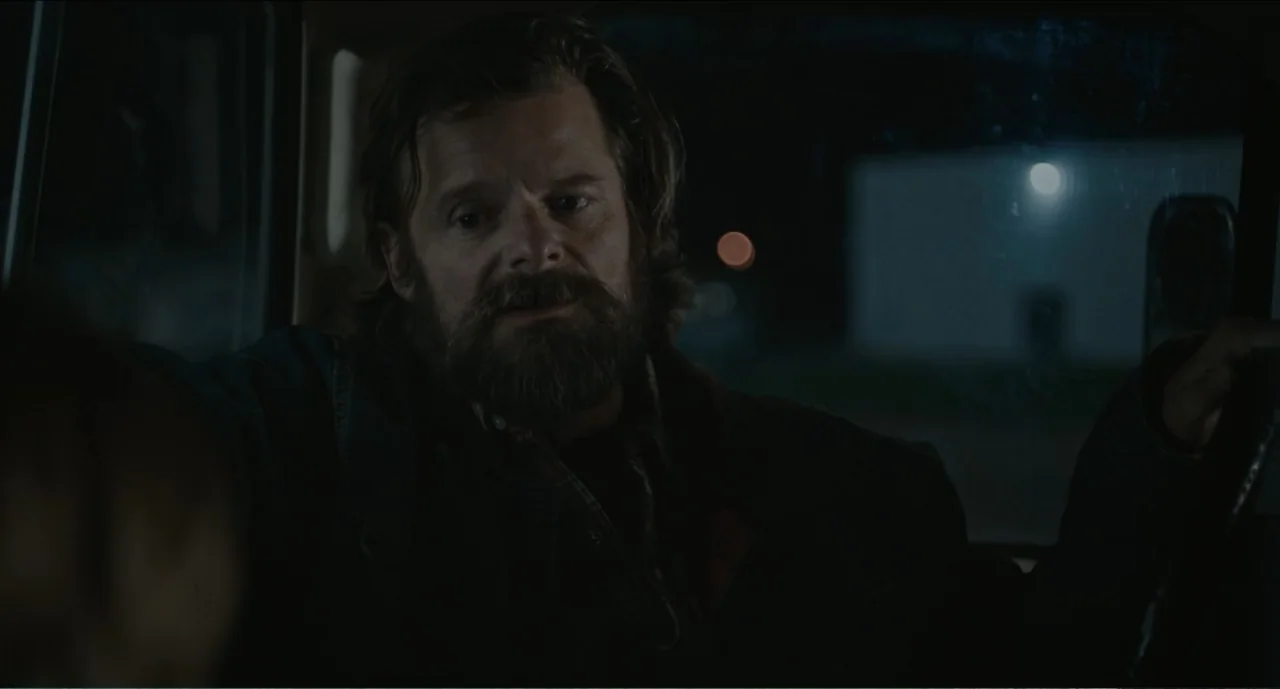 Cowboys (2014) movie still: medium — The interior of a vehicle at night with dim, low-contrast illumination. A man with a full beard sits in the…