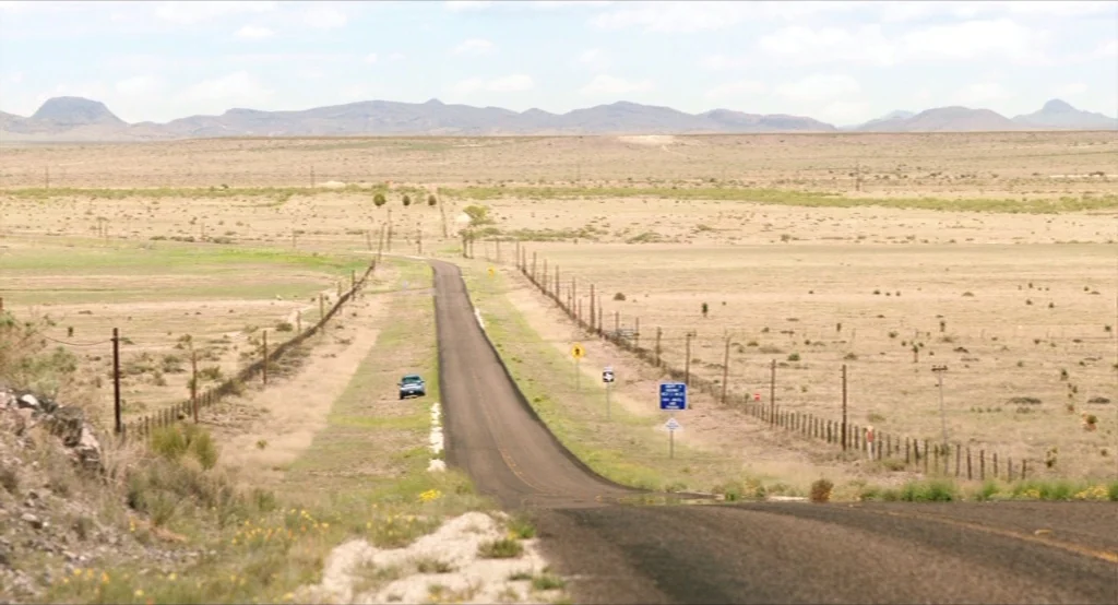 Results (2015) movie still: establishing — A vast, dry desert landscape stretches toward distant, low-lying mountains under a pale sky. A narrow…