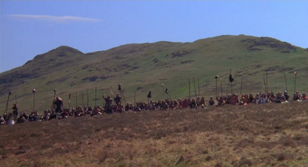 Monty Python and the Holy Grail (1975) movie still: wide — A vast, rolling green highland landscape slopes upward toward a distant ridgeline. A…