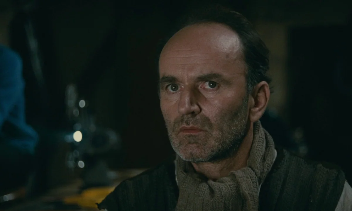 Muriel (1963) movie still: closeup — A dark, dimly lit basement room with a blurred mechanical object in the background. An older man with stubble…