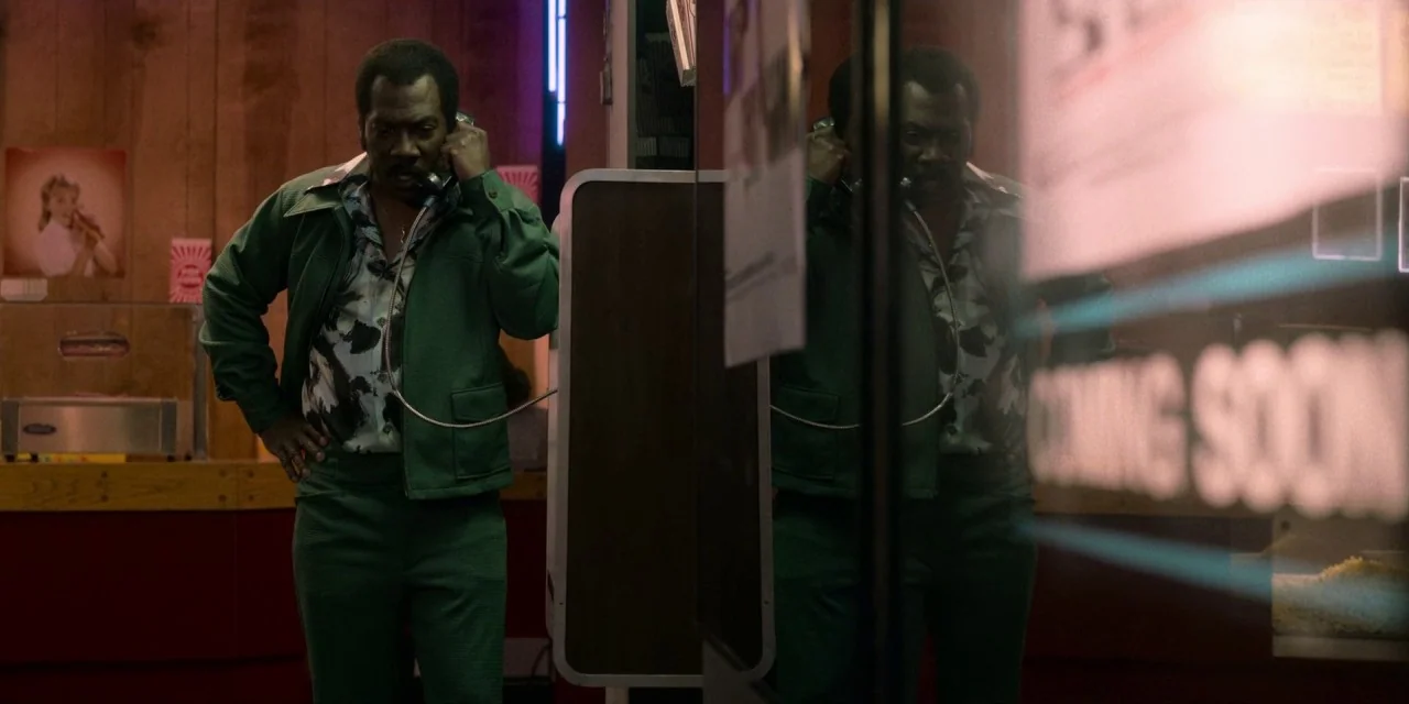 Dolemite Is My Name movie still: medium — A dimly lit lobby features wood-paneled walls and a reflection in a glass partition. A man wearing a green…