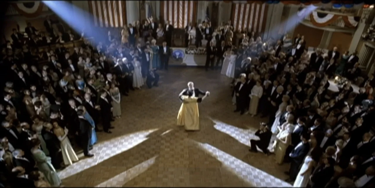 Path to War (2003) movie still: wide — A large ornate ballroom filled with guests in formal attire sits under dramatic spotlights. A single couple…