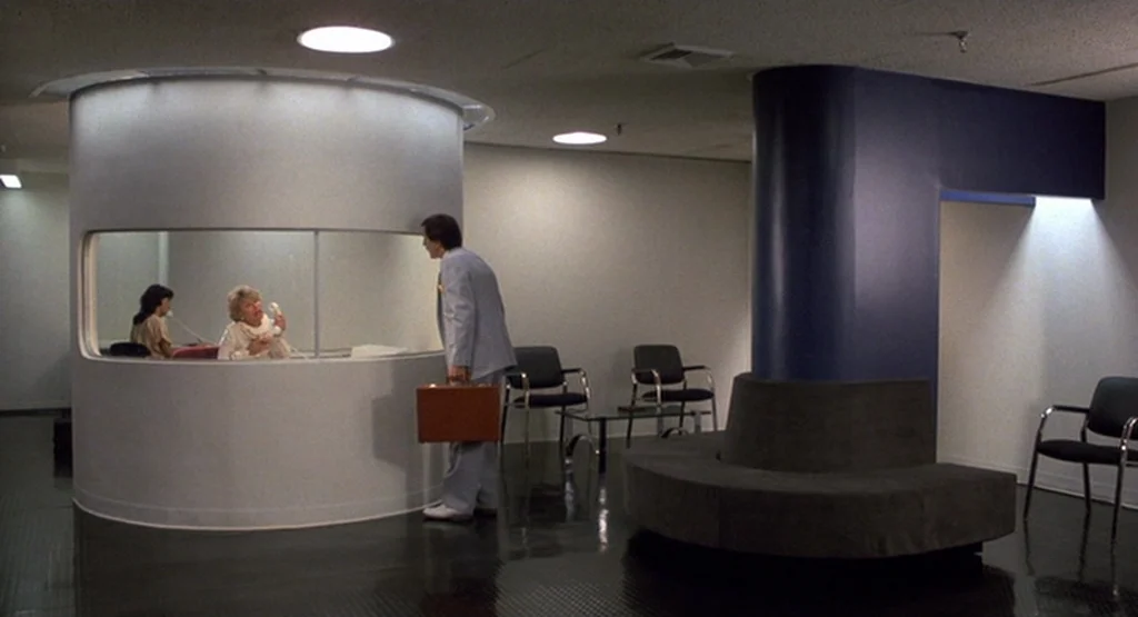 The King Of Comedy (1982) movie still: wide — A sterile reception area features a curved central desk and a dark curved bench. A man holding a brown…
