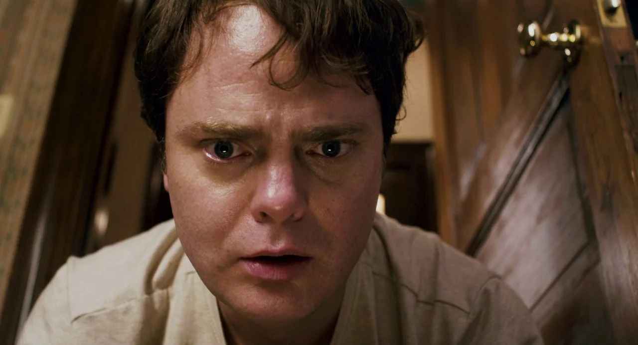 Super (2010) movie still: closeup — A man looks down at the camera from a high angle inside a home. The background shows wooden door trim and…