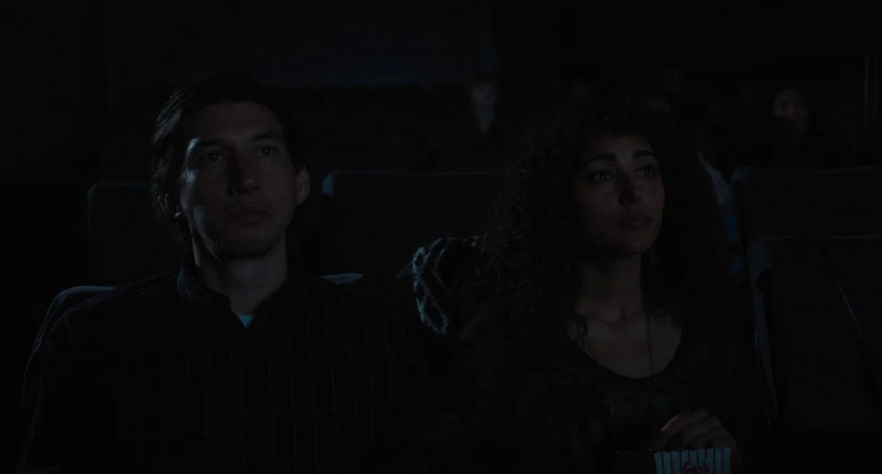 Paterson (2016) movie still: medium — Two individuals sit in dark upholstered chairs inside a dimly lit movie theater space. The characters look…