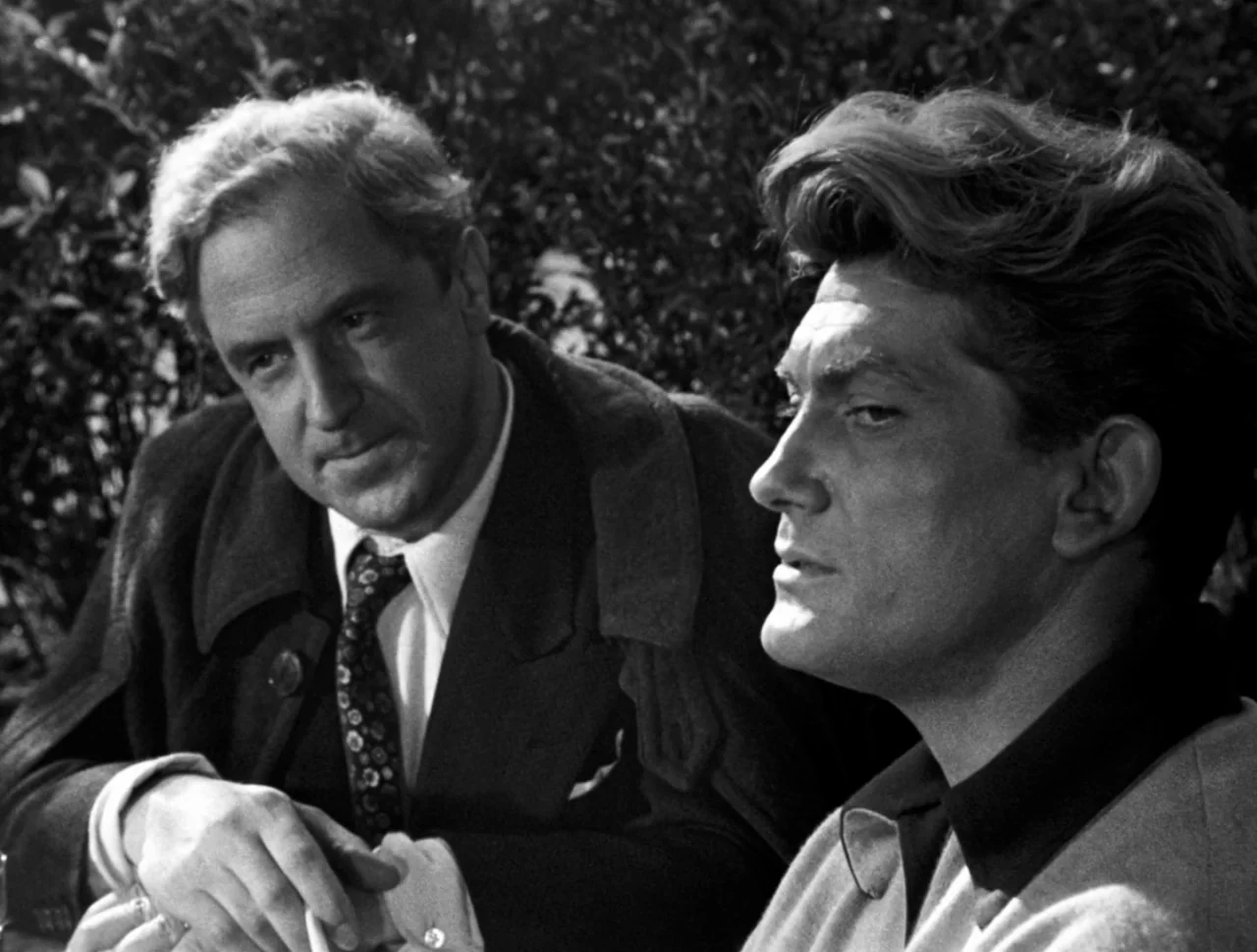 Orpheus (1950) movie still: medium — An outdoor area with thick green foliage blurred in the background. Two men sit together while facing slightly…