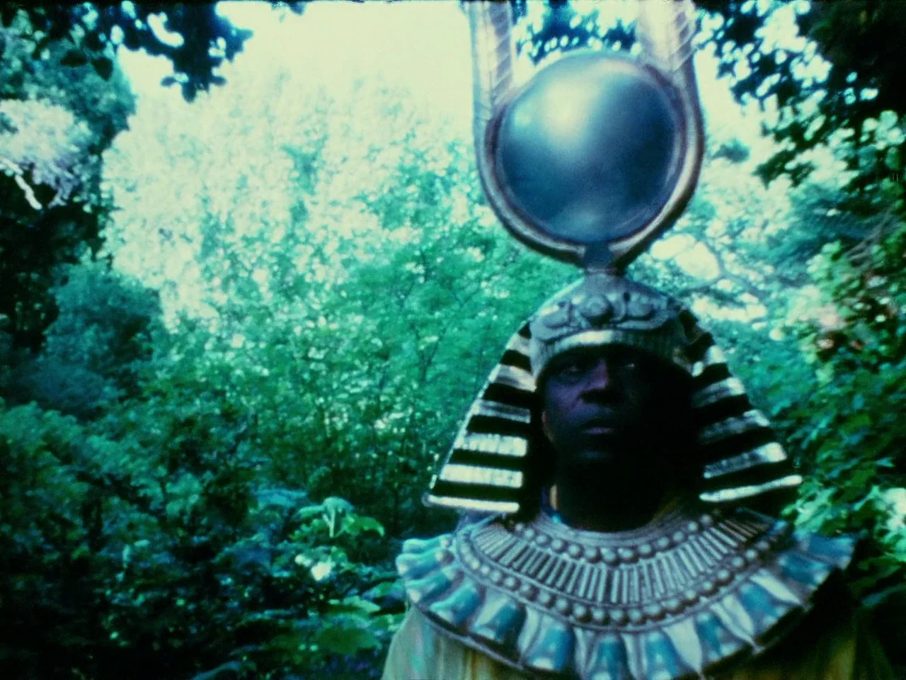 Space is the Place (1974) movie still: medium — A person wearing an elaborate Egyptian-style costume stands amidst a dense, green leafy forest. They…