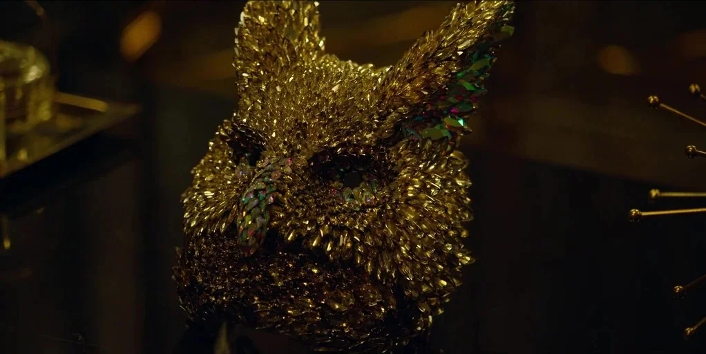Making of: Squid Game (2021) movie still: closeup — A close-up of an owl-shaped mask covered in gold and iridescent crystals. The object rests on a…