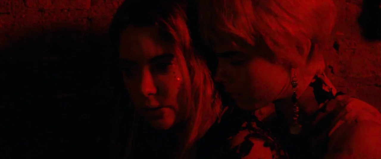 Her Smell (2018) movie still: closeup — A dark indoor space is bathed in intense red light against a textured wall. Two people stand close together…