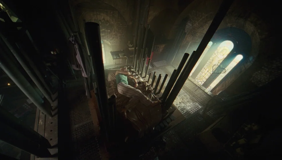 The Zero Theorem (2013) movie still: wide — An interior space within a bell tower featuring large metal pipes and stone architecture. A lone man…