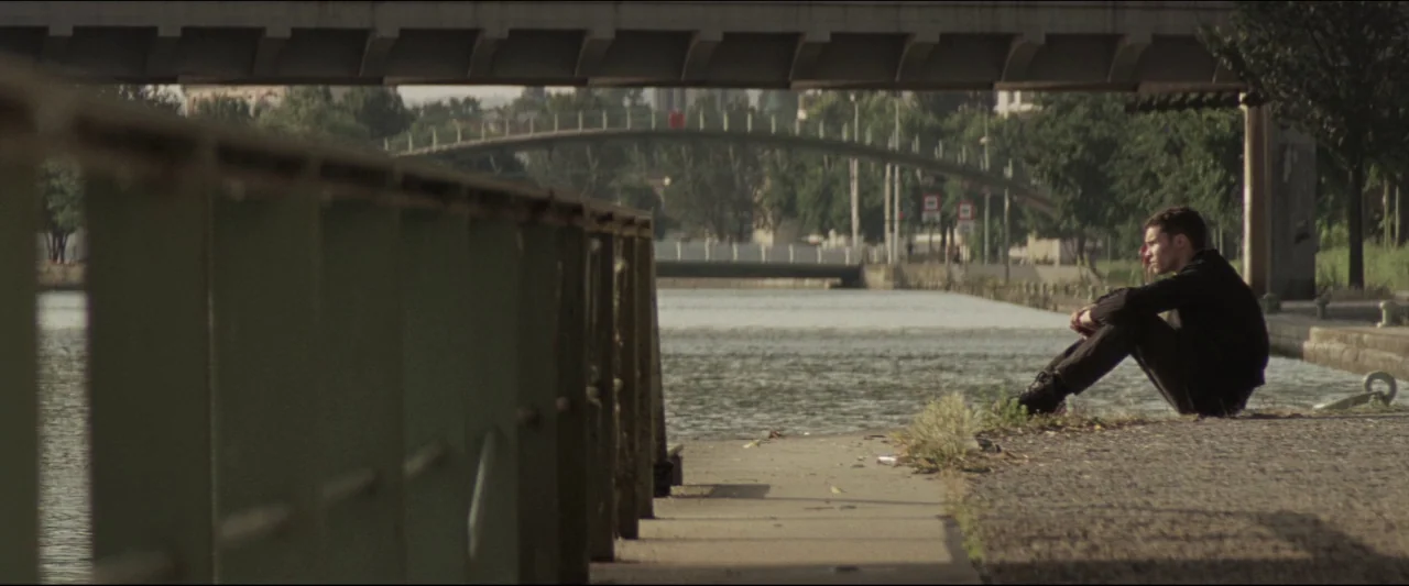 Colt 45 (2014) movie still: wide — A man sits on a concrete path alongside a water channel beneath a bridge. Trees and a distant pedestrian bridge…