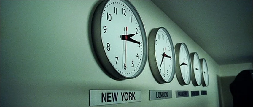 25th Hour (2002) movie still: medium — A series of round wall clocks with location labels are mounted on a wall. The clocks display varying times…