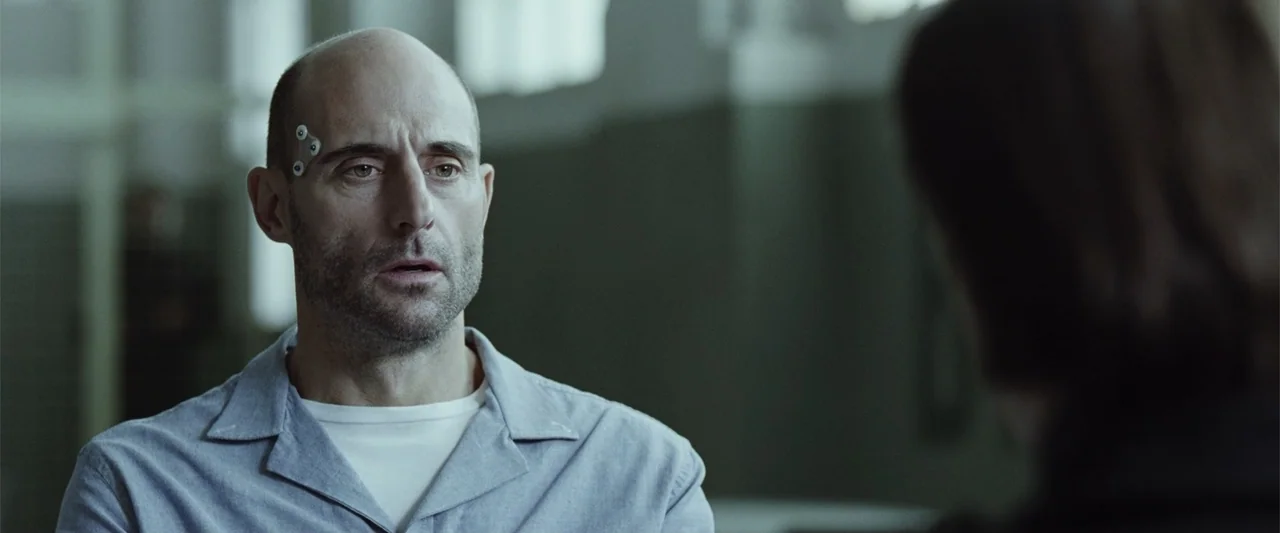 Anna (2013) movie still: medium — An interior space with a muted green wall and blurred institutional background elements. A middle-aged man wearing…