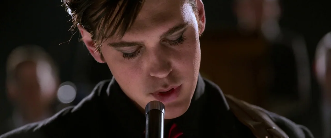 Elvis (2022) movie still: closeup — A dark concert stage focuses on the face of a young male performer. He is singing into a silver microphone with…