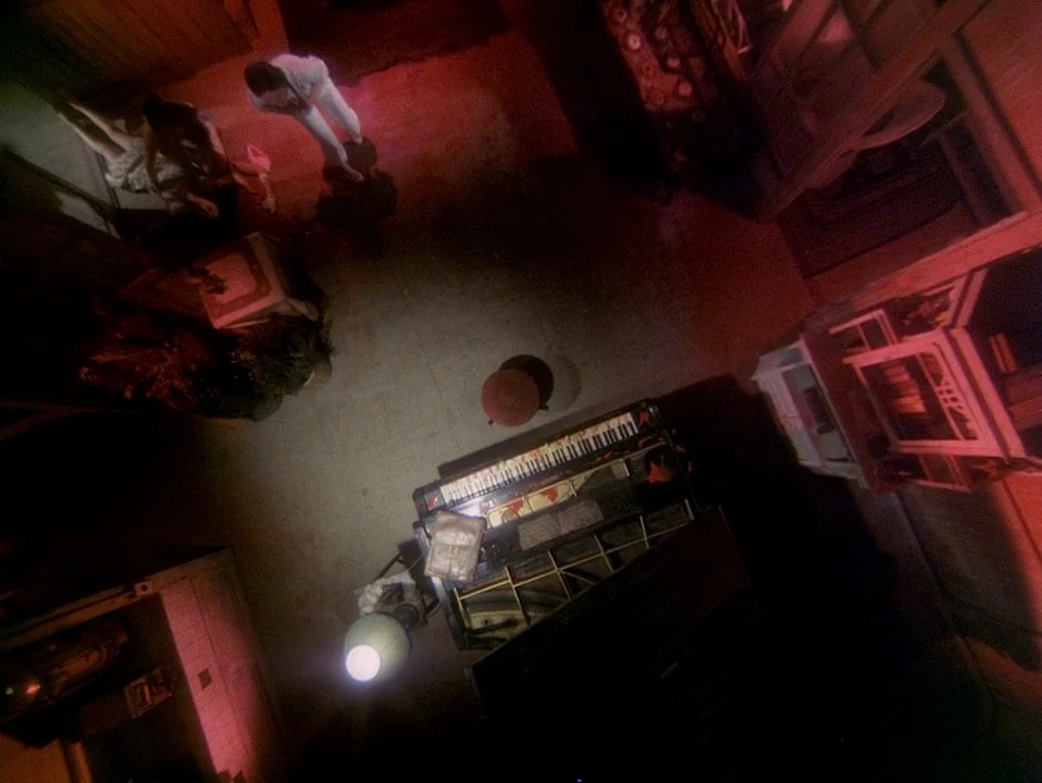 House (Hausu) (1977) movie still: wide — A top-down view shows a room with a grand piano and a floor lamp. Two figures stand together in the top…