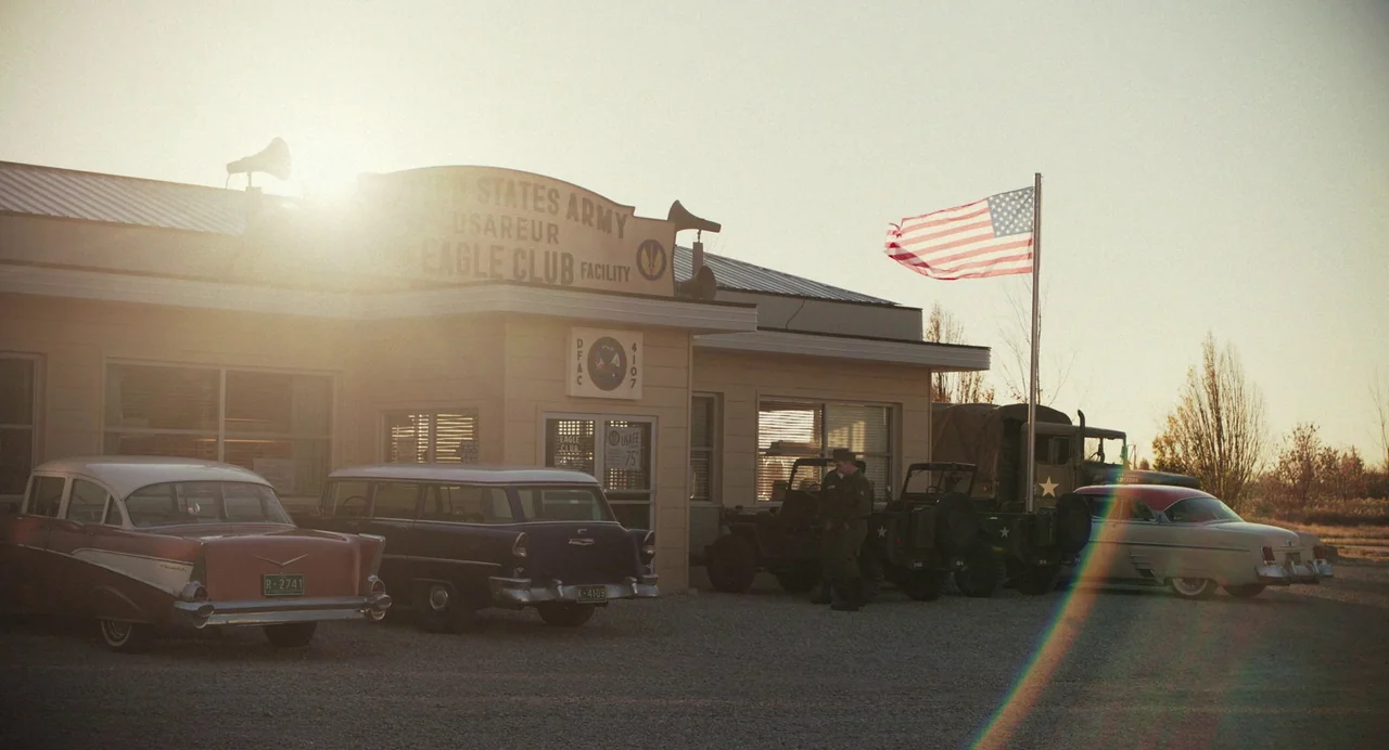 Priscilla (2023) movie still: wide — A single-story United States Army facility with a flag flying in the golden sunlight. Several mid-century…