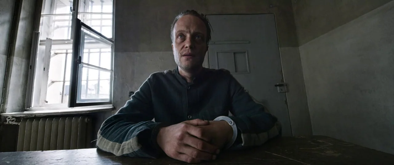 A Hidden Life (2019) movie still: medium — A dimly lit room features a barred window and a solid wooden door. A man sits at a table with his…
