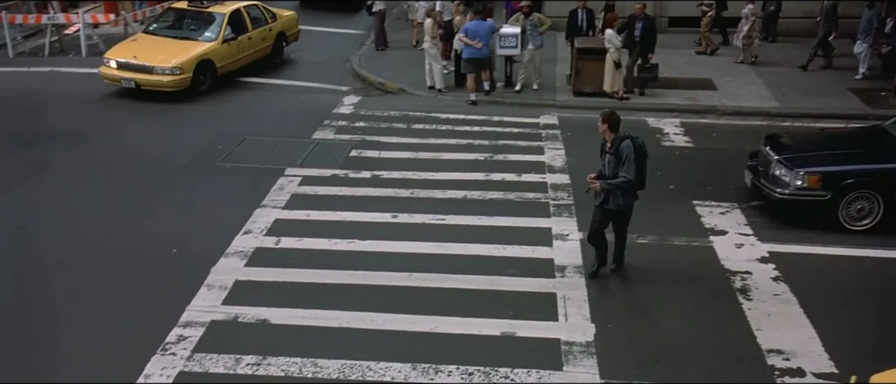 The Peacemaker (1997) movie still: wide — An urban intersection features a white striped pedestrian crosswalk on a dark asphalt road. A man in a…