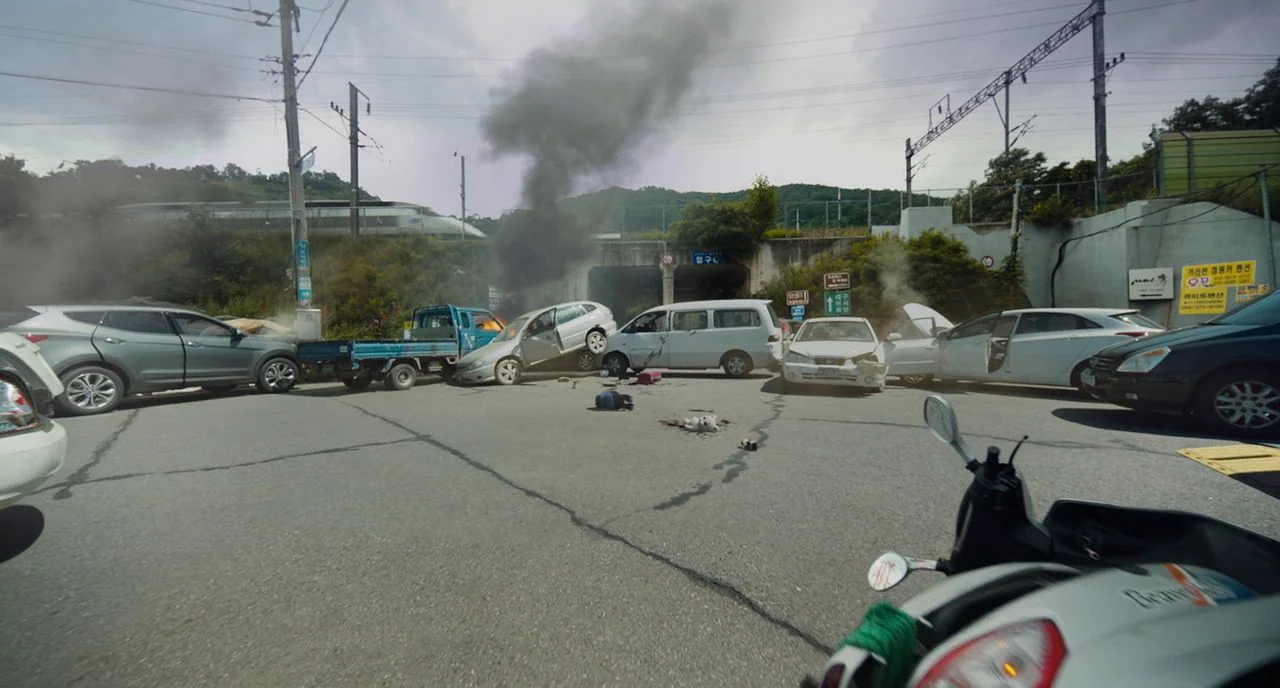 Train To Busan movie still: wide — An intersection serves as the site of a multi-car pileup with significant structural damage. Vehicles are…