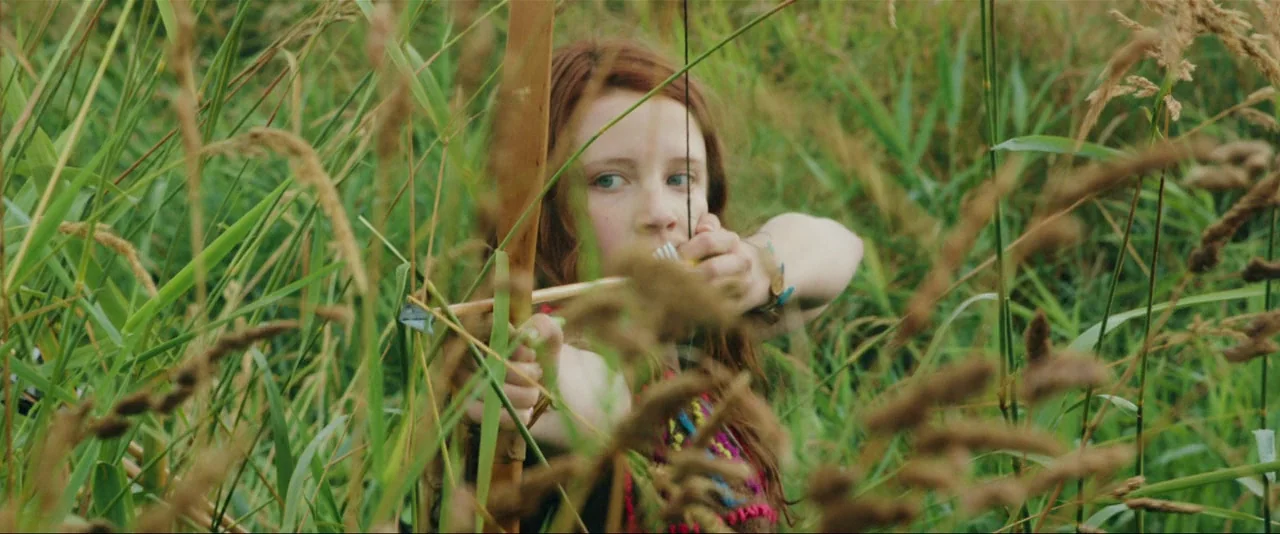 Captain Fantastic (2016) movie still: medium — A young girl with auburn hair hides within a dense field of tall grass. She pulls back the string of…