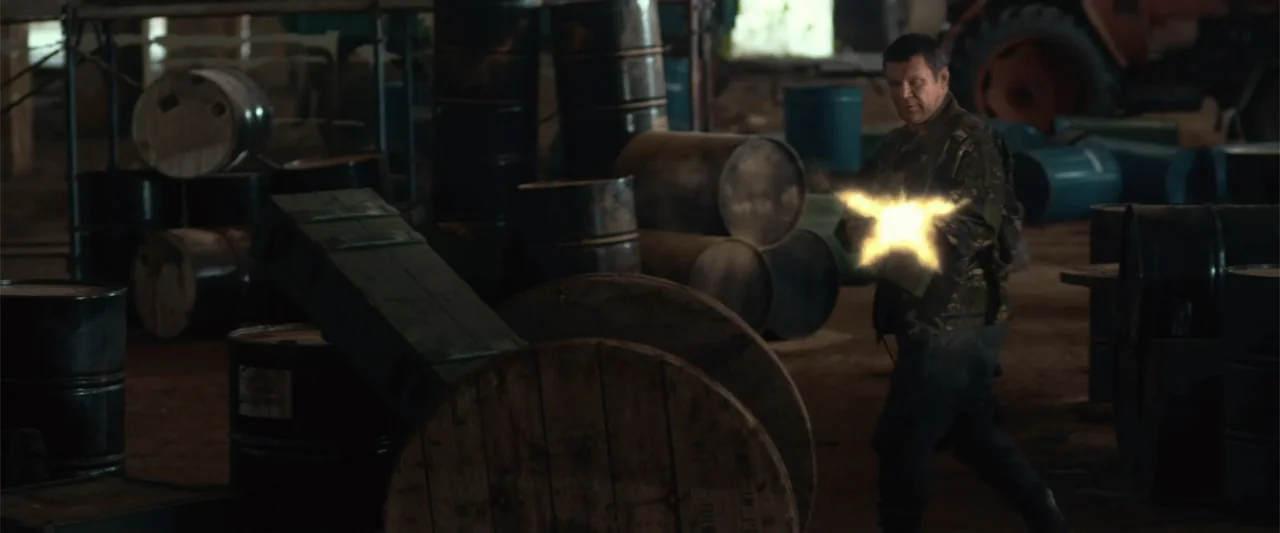 The Condemned 2 (2015) movie still: medium — A dark industrial space contains stacks of metal drums and large wooden spools. An older man in…
