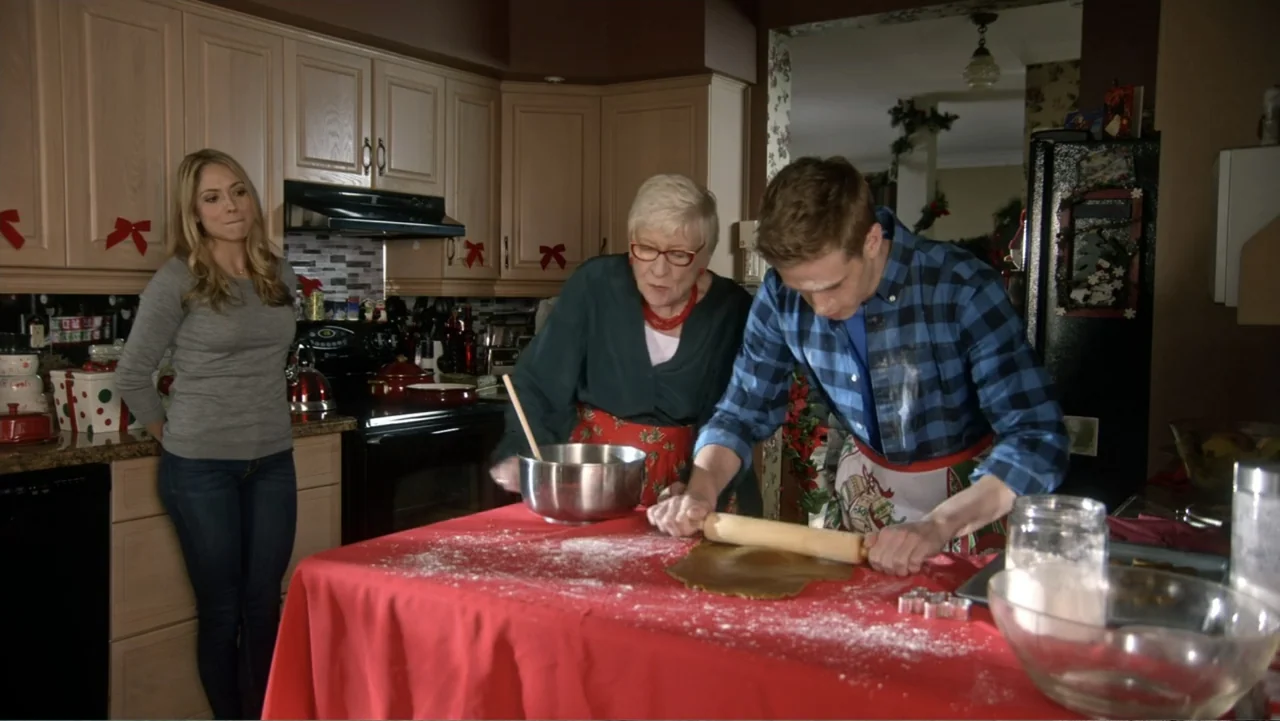 The Christmas Cure (2017) movie still: medium — A residential kitchen with light wood cabinets and a red holiday tablecloth. A man rolls out dough…