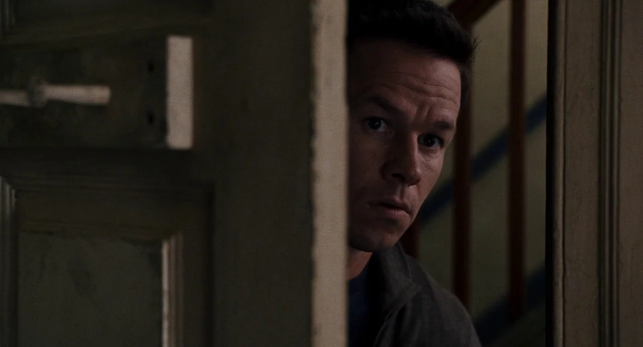 The Happening (2008) movie still: closeup — A man peers through a partially opened door in a dimly lit, dark space. The subject stands in the gap…