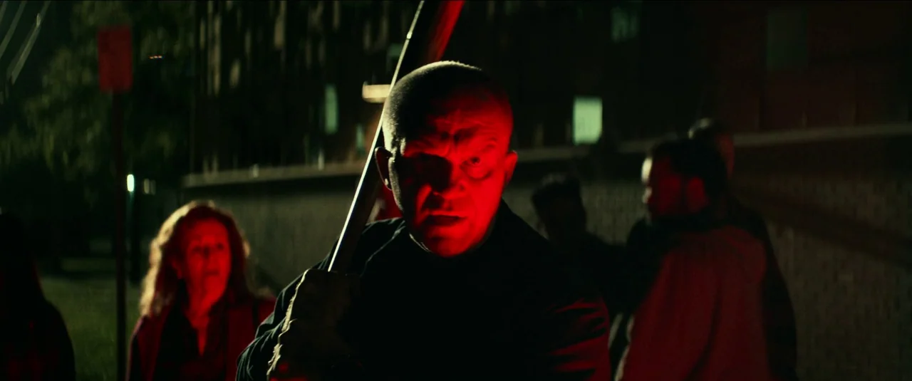 Halloween Kills (2021) movie still: medium — An outdoor night scene in a dimly lit space with dark brick walls. A bald man holds a baseball bat…