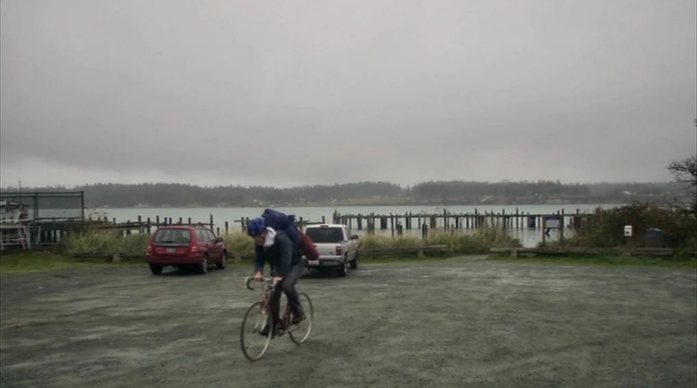 Your Sister’s Sister (2011) movie still: wide — An overcast waterfront parking lot with a gravel surface and two parked cars. A cyclist rides across…