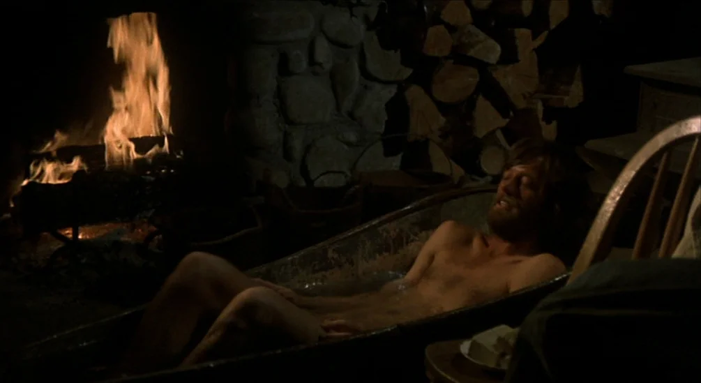 The Hired Hand (1971) movie still: medium — A dimly lit room features a metal bathtub positioned in front of a fireplace. A man with a beard rests…