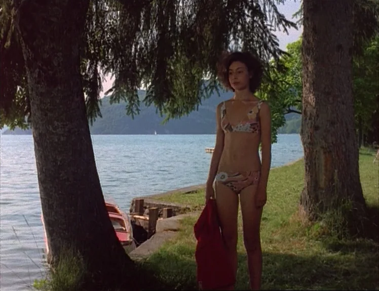 Claire’s Knee (1970) movie still: fullbody — A woman stands on a grassy bank beside a large lake surrounded by trees. She is positioned between two…