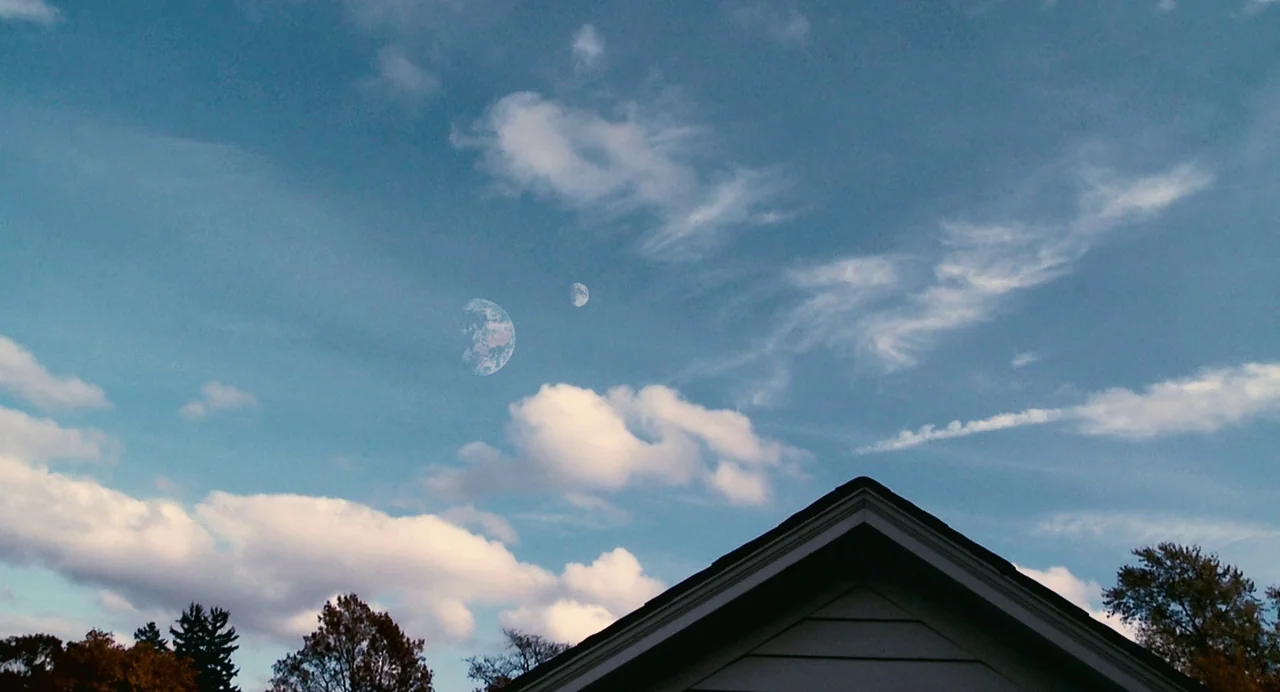 Another Earth (2011) movie still: establishing — A residential rooftop edge frames a clear daytime sky with scattered white clouds. The planet Earth…