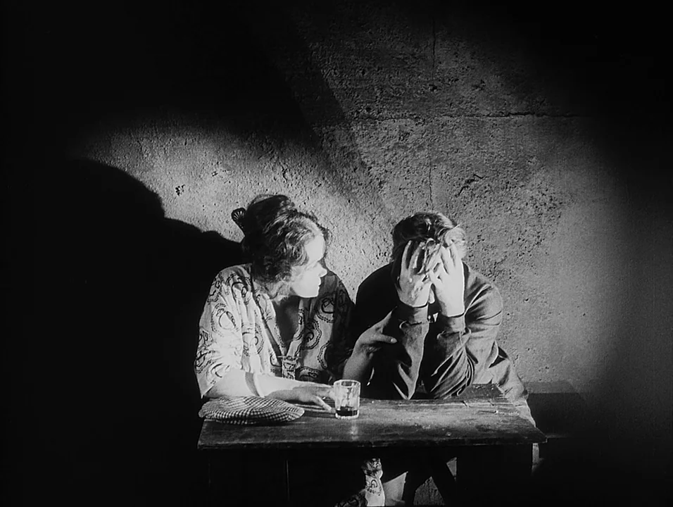 Coeur Fidele (1923) movie still: medium — Two people sit at a small wooden table against a dark, textured concrete wall. The man covers his face…