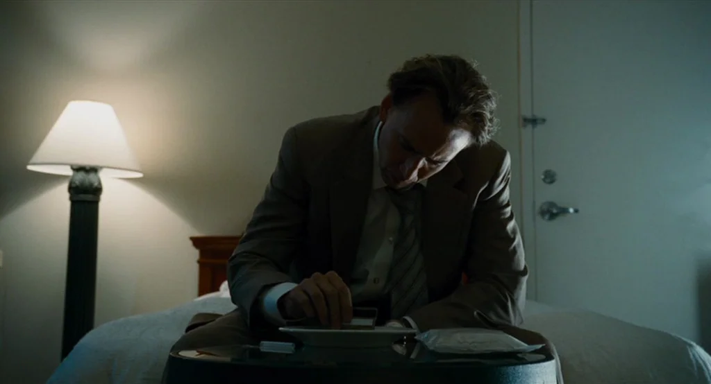 Bad Lieutenant: Port Of Call New Orleans (2009) movie still: medium — A dim motel room features a beige wall and a simple door frame. A man sits on…