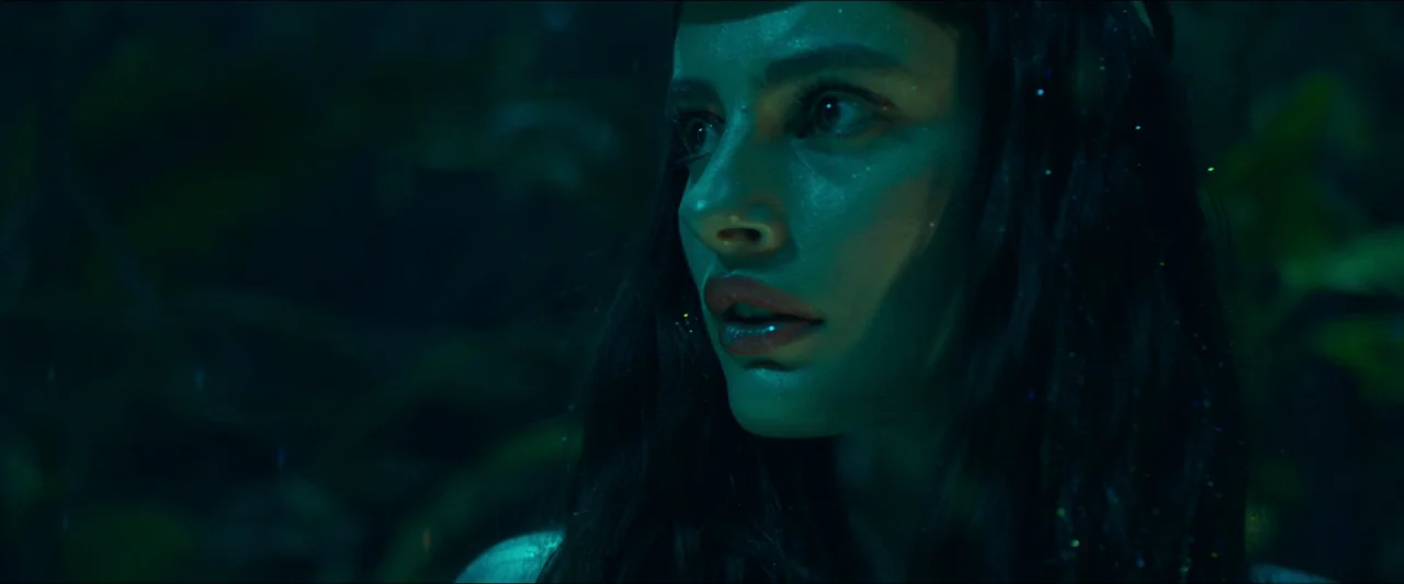 Birds of Paradise (2021) movie still: closeup — A dense dark environment filled with foliage exists under low-light conditions. A woman with long…