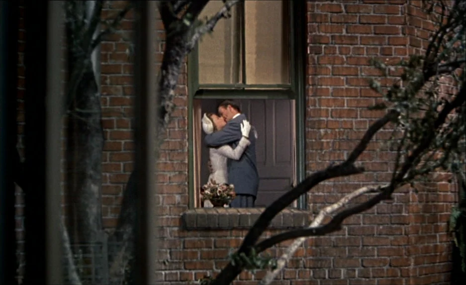 Rear Window (1954) movie still: medium — The exterior of a dark red brick building is visible at night. A couple embraces and kisses inside a…