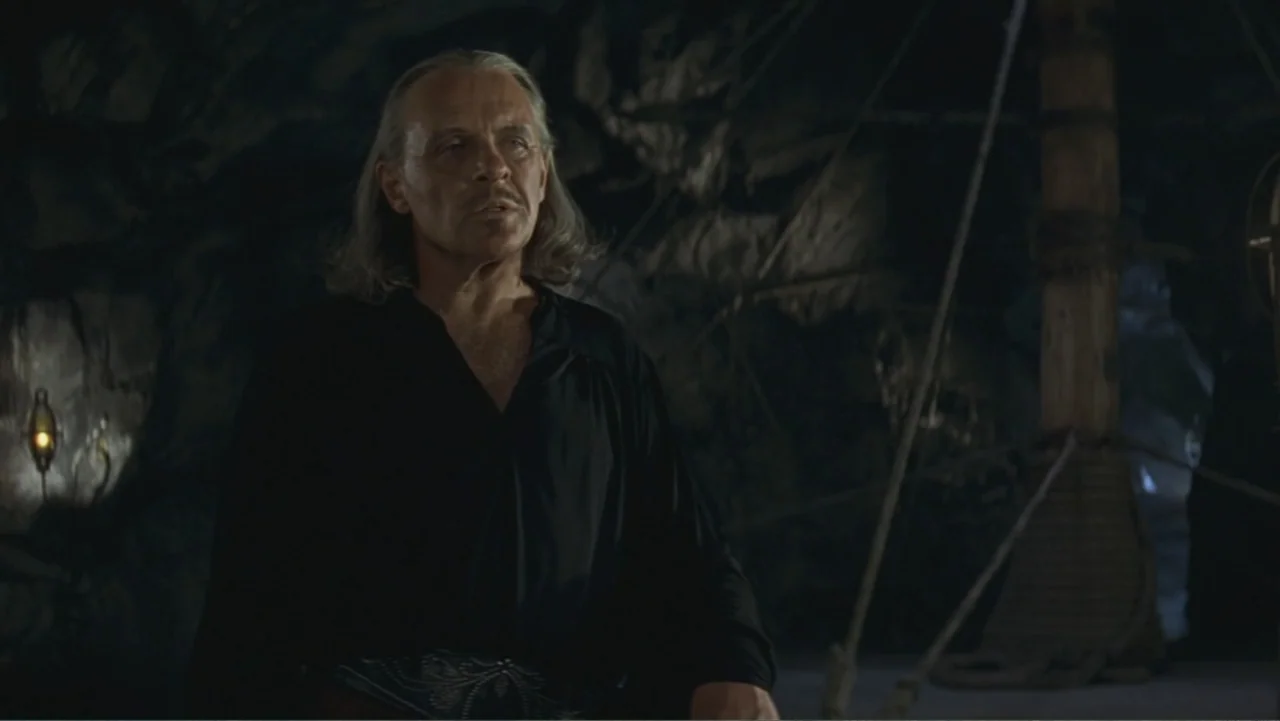 The Mask of Zorro (1998) movie still: medium — A dark, textured stone cave interior contains a singular dimly lit lamp. An older man with gray hair…