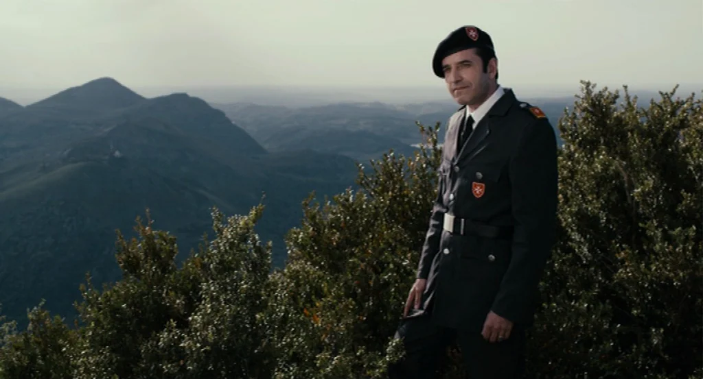 Lourdes movie still: medium — A man in a military uniform stands on a ridge overlooking a vast range. The mountain peaks stretch into the distance…